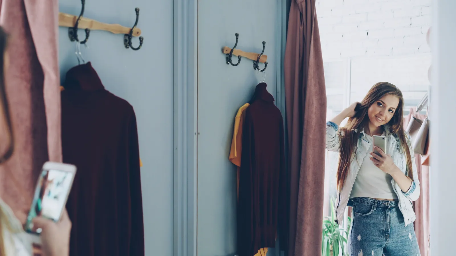 หญิงสาวยืนหน้ากระจกในห้องลองเสื้อ ถือมือถือถ่ายรูปตัวเอง มีเสื้อผ้าแขวนอยู่บนตะขอข้างหลัง