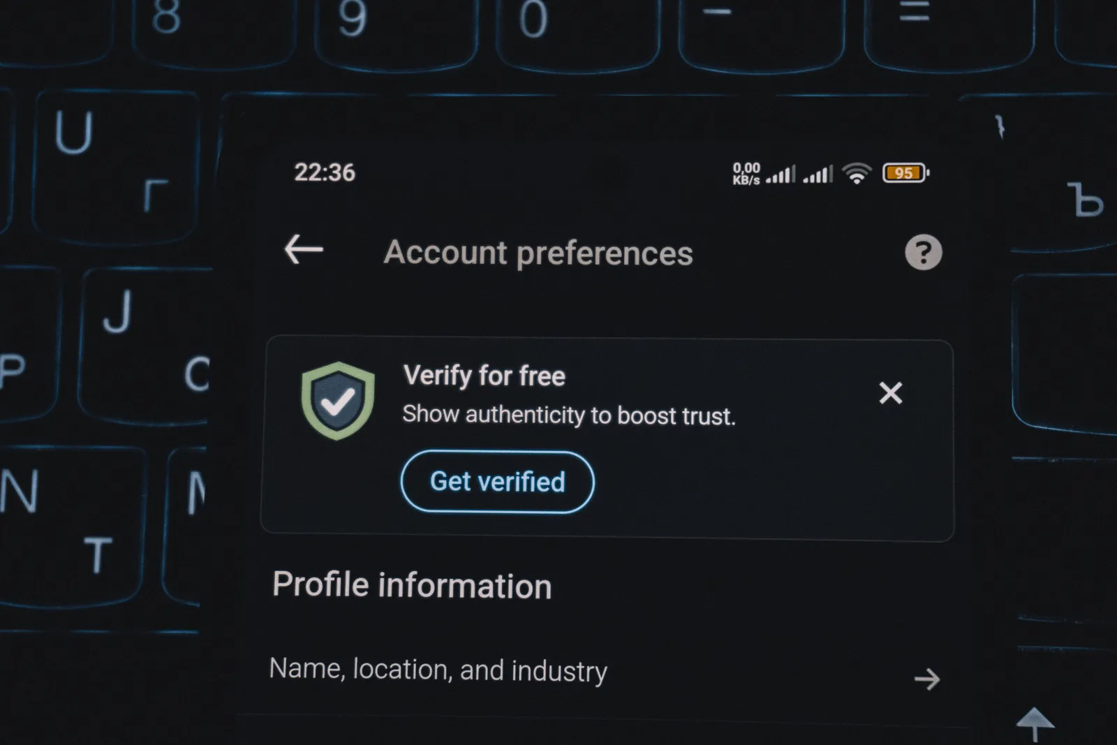 หน้าจอมือถือโหมดมืดแสดงหน้า Account preferences พร้อมแถบ Verify for free และปุ่ม Get verified วางอยู่บนคีย์บอร์ด